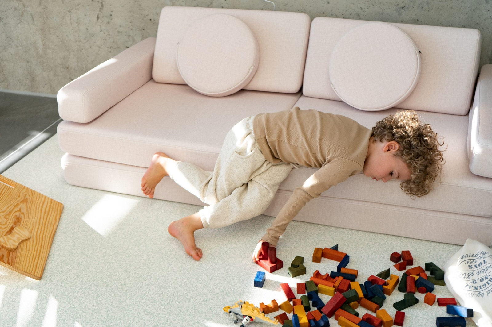Boy lying across the seat of a light pink play sofa, with coloured building blocks on the rug