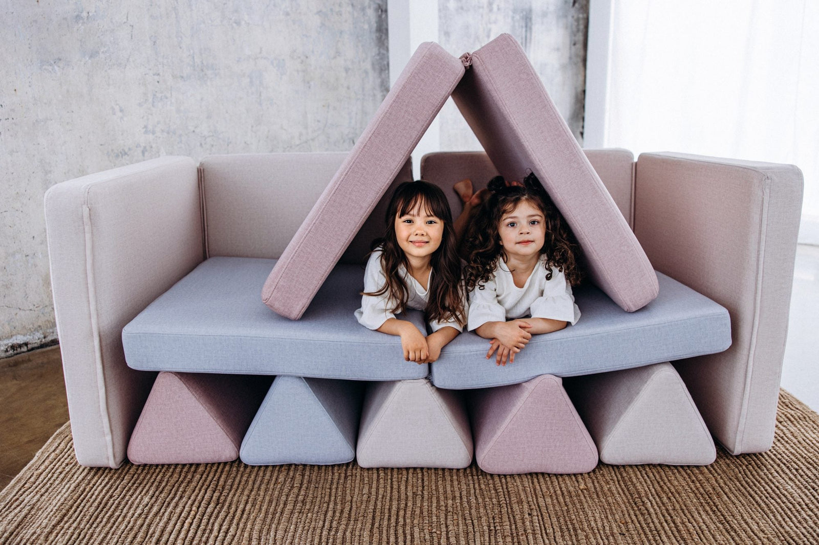 Two girls lying under a roof built from pink play sofa cushions