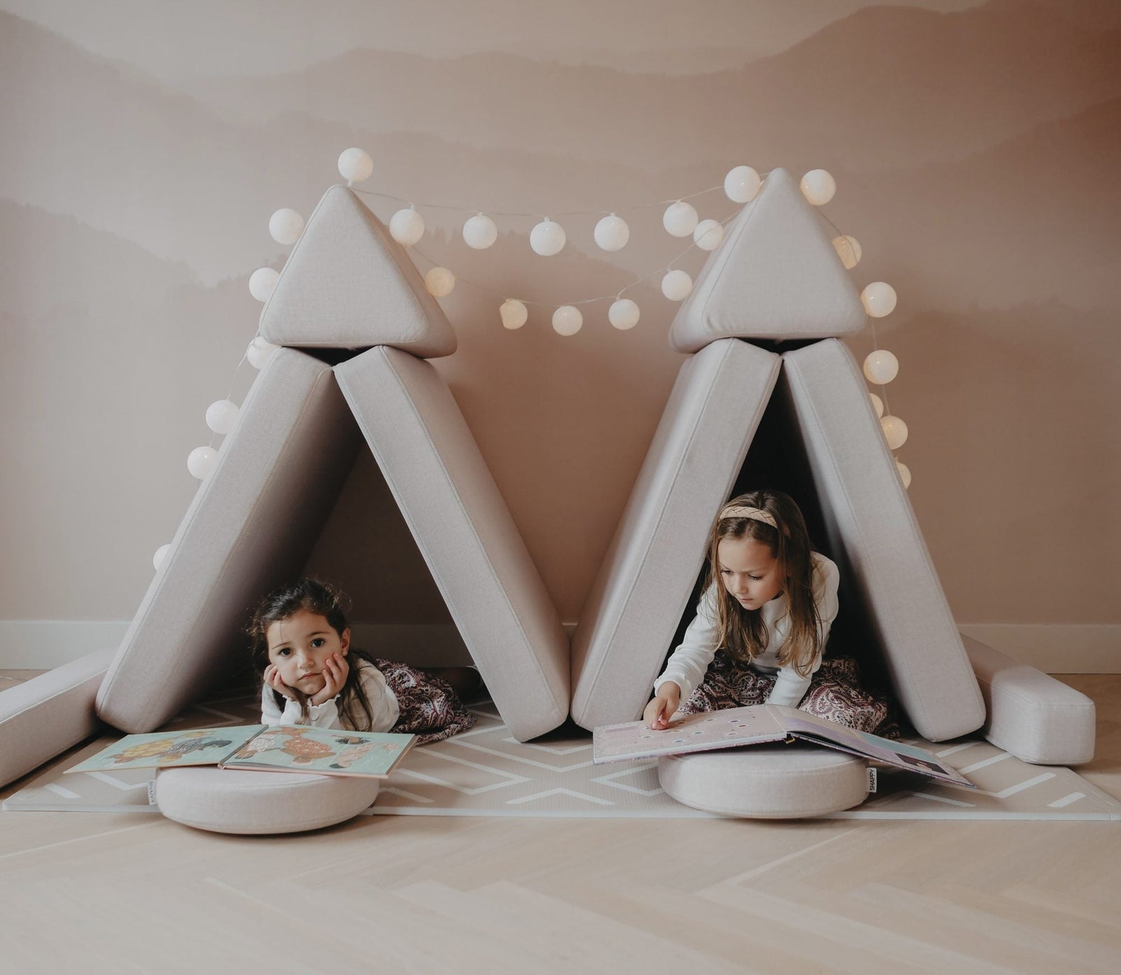 Two girls reading in two tents built from two play sofas, with a string of lights