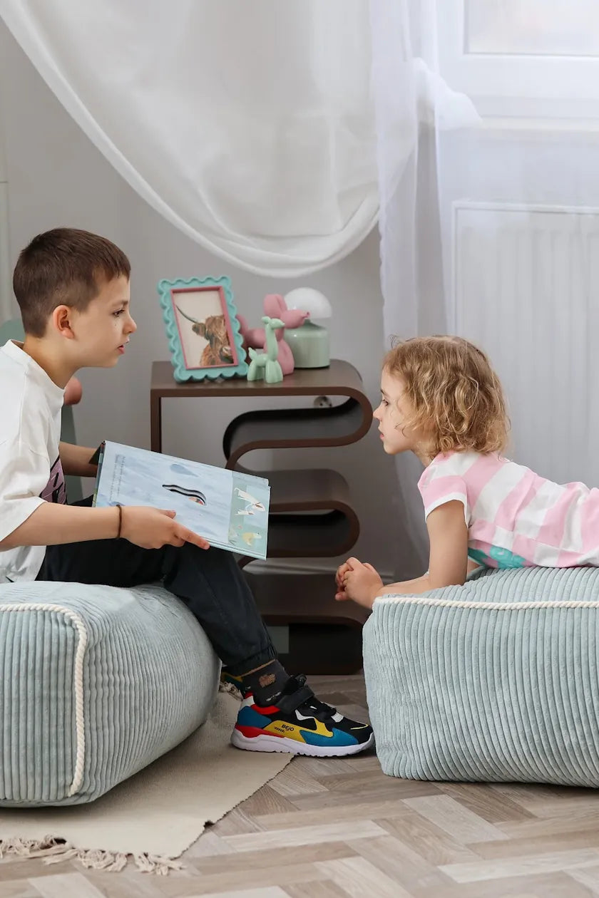 Boy sitting on Kumo Ottoman Peppermint while reading to his sister