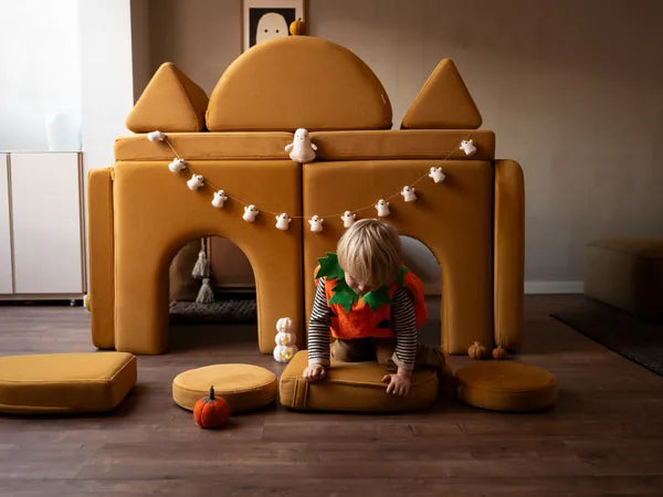 Garçon déguisé en citrouille sortant d’un château d’Halloween fait d’arches Shappy et d’éléments du canapé de jeu, avec une guirlande de fantômes
